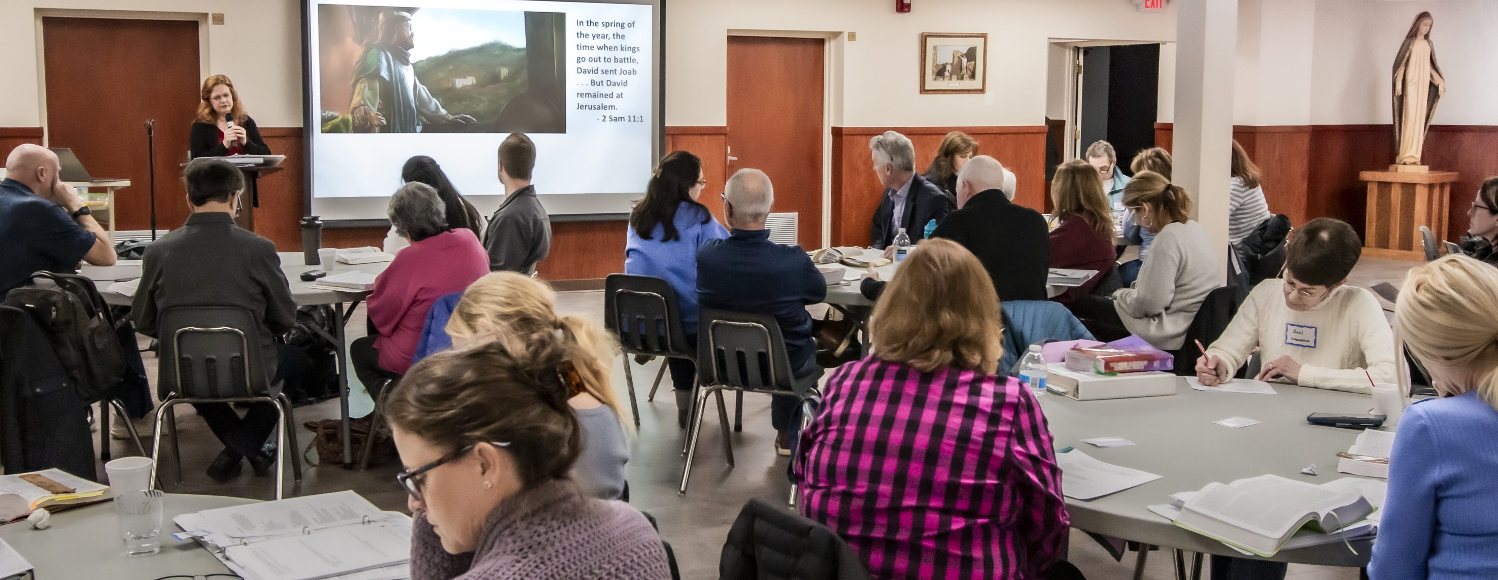 A live, in-person speaker teaches the Bible to a room of adults at circle tables.